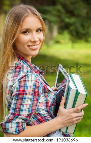Student outdoors. Cheerful young female student holding books and digital tablet while standing in a park