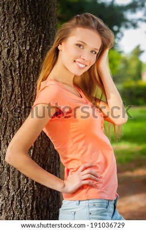 Beauty in nature. Side view of beautiful young woman holding hand in hair and smiling while leaning at the tree