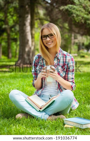 Searching inspiration outdoors. Beautiful young female student adjusting her glasses and smiling while sitting in a park with books around her