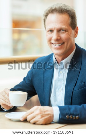 Having a break with fresh coffee. Cheerful mature man in formalwear drinking coffee and smiling while sitting in coffee shop