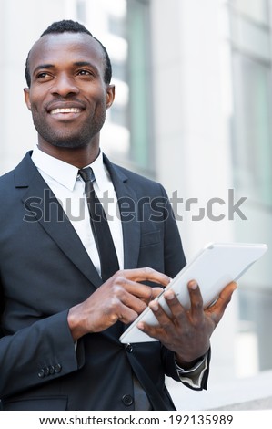 Businessman with digital tablet. Confident young African man in formal wear working on digital tablet and looking away with smile while standing outdoors