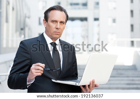 Business expert. Confident senior man in formal wear holding laptop and looking at camera while standing outdoors
