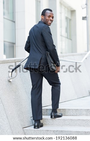 Cheerful businessman. Cheerful young African man in formal wear moving up by staircase and looking over shoulder