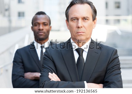 Confident business experts. Confident mature man in formal wear keeping arms crossed and looking at camera while young African man standing behind him