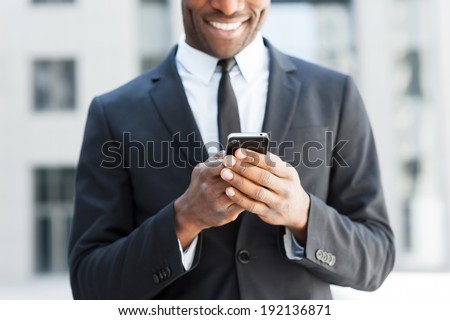 He is always in touch. Cropped image of cheerful young African man in formal wear holding mobile phone and smiling while standing outdoors