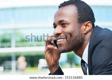 Businessman on the phone. Side view of handsome young African man in formal wear talking on the mobile phone and smiling while standing outdoors