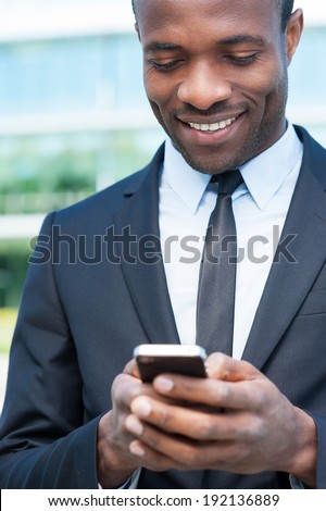 Typing business message. Cheerful young African man in formal wear holding mobile phone and smiling while standing outdoors