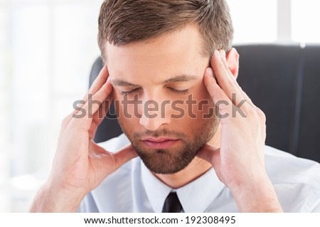 Feeling tired and depressed. Depressed young man in shirt and tie holding head in hands and keeping eyes closed while sitting at his working place