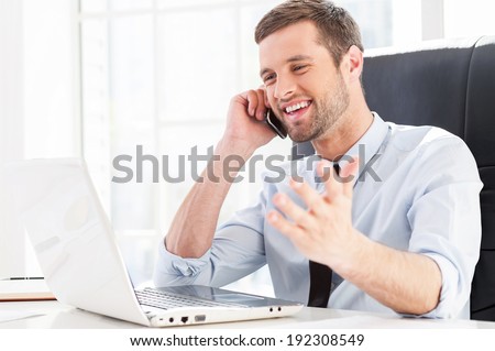 Happy with business going. Happy young man in formal wear looking at computer monitor and talking on the mobile phone while sitting at his working place
