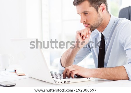 Concentrated on work. Side view of thoughtful young man in formal wear looking at laptop and holding hand on chin while sitting at his working place