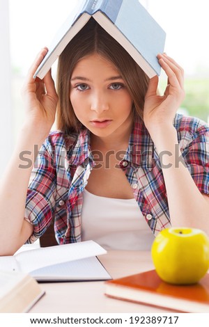 Feeling tired of studying. Frustrated teenage girl carrying book on head and looking at camera while sitting at the desk with books laying on it