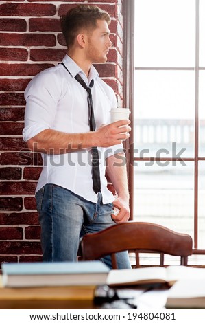 Refreshing his mind. Handsome young man in shirt and tie holding coffee cup and looking through the window while sitting at the window sill