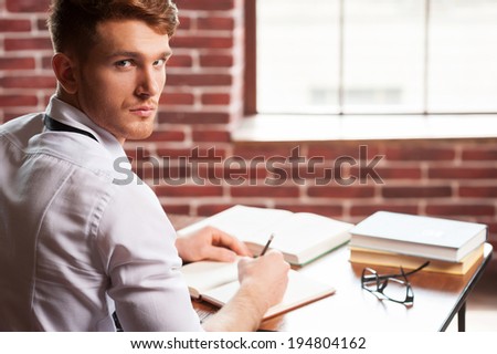 Confident author. Handsome young man in shirt and tie writing something in note pad and looking over shoulder while sitting at his working place