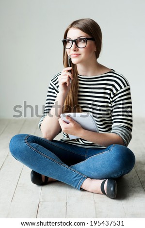 Waiting for inspiration. Thoughtful young woman in striped clothing holding note pad and touching her chin with pen while sitting on the hardwood floor