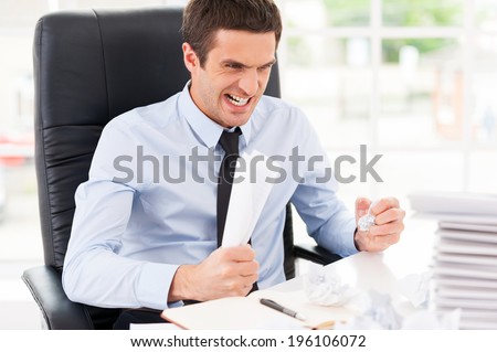 Sick and tired. Furious young man in shirt and tie holding papers in hands and shouting while sitting at his working place