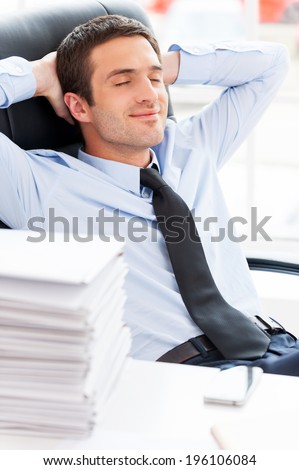 Businessman day dreaming. Handsome young beard man in shirt and tie holding hands behind head and smiling while sitting at the table with stack of paperwork laying on it