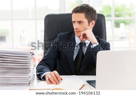 Missing the deadlines. Thoughtful young man in shirt and tie looking at the stack of paperwork and holding head on chin while sitting at his working place