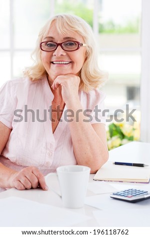 Getting finances straight. Happy senior woman holding hand on chin and smiling at camera while sitting at the table