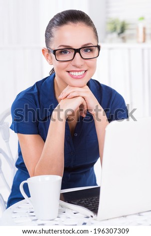 Smart and creative. Beautiful young woman holding hand on chin while sitting at the table with laptop and cup laying on it