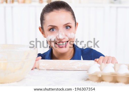Messy cooking. Playful young woman with flavor on face holding rolling pin and looking out of the kitchen table