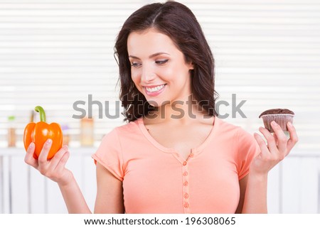 I choose healthy food. Cheerful young woman choosing what to eat while holding chocolate muffin and fresh pepper