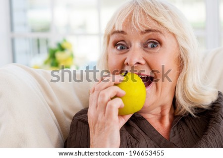 I love apples! Cheerful senior woman holding apple and looking at camera while sitting at the chair