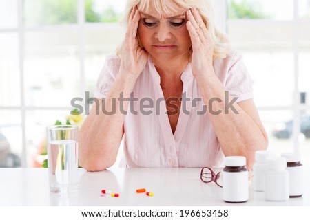 Feeling bad. Depressed senior woman holding head in hands and looking at the pills laying on the table