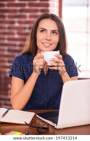 Dreaming up business ideas. Thoughtful young woman holding cup with hot drink and looking away while sitting at her working place