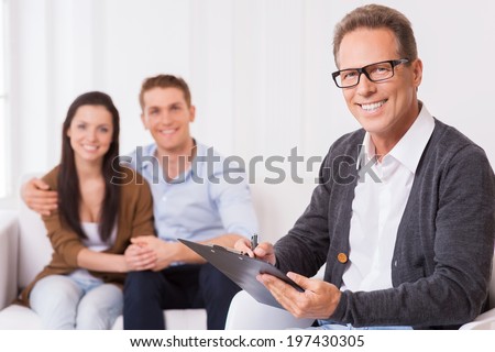 Confident family psychologist. Confident psychiatrist writing something in clipboard and smiling while cheerful couple sitting in the background and holding hands