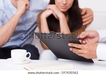 Sign here. Close-up of thoughtful young couple sitting on the couch while man in formalwear pointing clipboard with document
