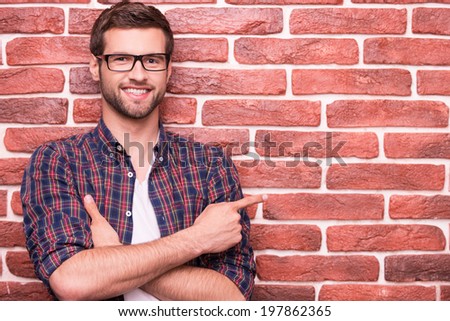 Confident handsome pointing your advertising. Handsome young man keeping arms crossed and pointing at copy space while standing against brick wall