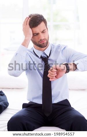 Late again. Frustrated young man in shirt and tie checking the time and touching head with hand while sitting on the bed