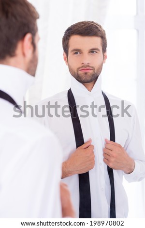 Preparing to a special day. Handsome young man in white shirt and untied necktie standing against mirror