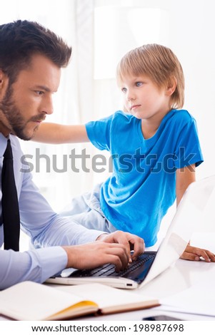 Play with me daddy! Busy young man in shirt and tie working on laptop while his son sitting close to him and touching his shoulder