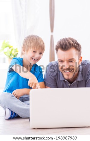Spending time together. Happy father and son surfing the net at home while little boy pointing laptop monitor and smiling