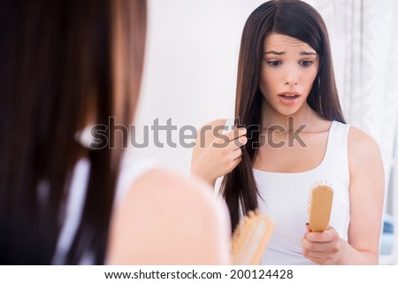 Hair loss. Depressed young woman looking at her hairbrush and expressing negativity while standing against mirror