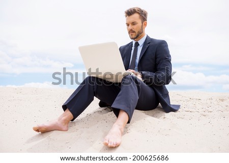 Businessman in desert. Confident young man in formalwear working on laptop while sitting on sand in desert