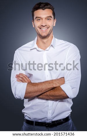 Confident and successful. Happy young man in white shirt looking at camera and keeping arms crossed while standing against grey background