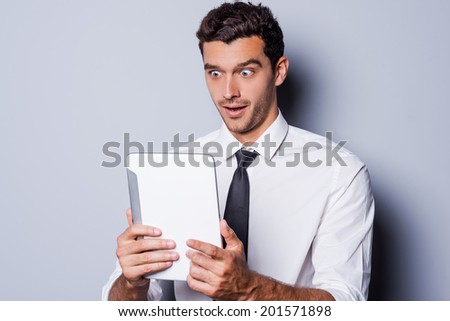 It is unbelievable! Surprised young man in shirt and tie looking at digital tablet and keeping mouth open while standing against grey background