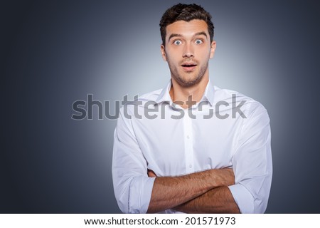 Wow! Surprised young man in white shirt staring at camera and keeping arms crossed while standing against grey background