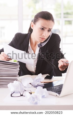 Busy office life. Frustrated young woman in formalwear talking on the telephone and holding documents while sitting at her working place