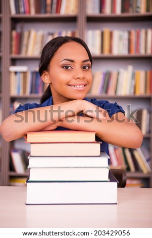 She loves studying. Confident young black woman leaning at the book stack and smiling while sitting at the library desk