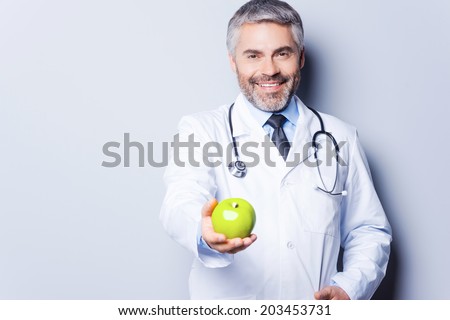 Eat healthy! Cheerful mature doctor in giving a green apple to you and smiling while standing against grey background