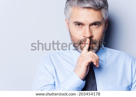 Keep my secret! Confident mature man in white shirt and tie looking at camera and holding finger on lips while standing against grey background
