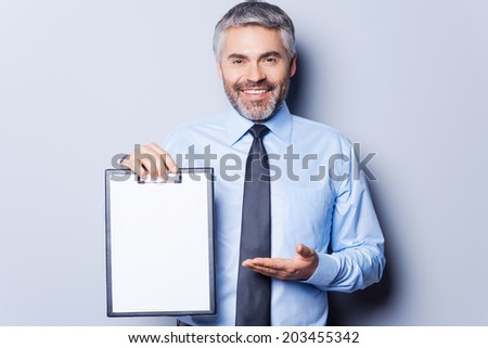 Your text here. Happy young man in formalwear holding clipboard and pointing it with smile while standing isolated on white background