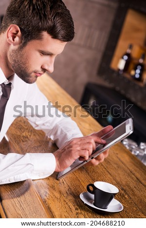 Staying in touch. Top view of confident young man in shirt and tie sitting at the bar counter and working on digital tablet