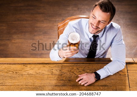 Enjoying his favorite beer. Top view of handsome young man in shirt and tie examining glass with beer and smiling while sitting at the bar counter
