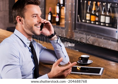 Good talk with friend. Side view of happy young man in shirt and tie talking on the mobile phone and gesturing while sitting at the bar counter