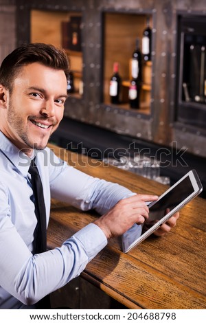 Surfing the net in bar. Handsome young man in shirt and tie working on digital tablet and smiling while sitting at the bar counter