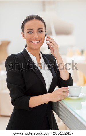 Enjoying coffee break. Beautiful young woman in formalwear drinking coffee and talking on the mobile phone while leaning at bar counter